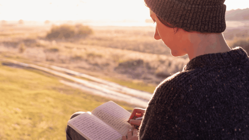 O impacto das palavras na alma humana Uma mulher está sentada ao ar livre, escrevendo em um diário ou caderno, com um pôr do sol ao fundo.
