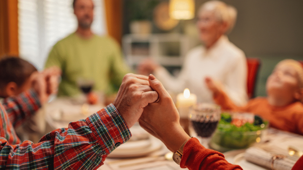 Duas mãos dadas numa mesa de refeição, ao fundo, uma família reunida, com adultos e crianças em clima acolhedor.