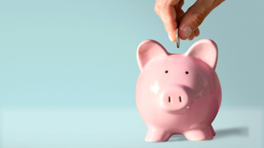 Rituais da Lua para atrair mais dinheiro mão colocando uma moeda em um cofre de porquinho rosa em fundo azul