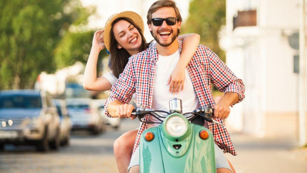 Mulher e homem numa moto, sorrindo e andando numa rua.